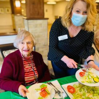 A caregiver serves a meal to an elderly woman at a dining area.