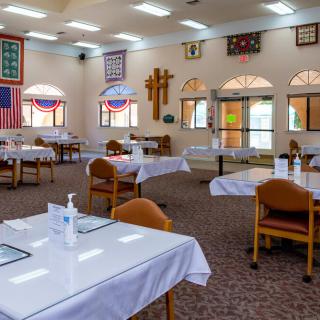 An empty dining hall with neatly arranged tables and chairs, featuring wall decorations and large windows.