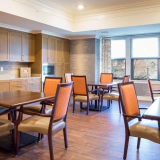 A cozy dining area with wooden tables and chairs, featuring a kitchen space and large windows.