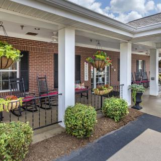 A cozy porch with rocking chairs, hanging plants, and a neatly landscaped area in front of a brick building.