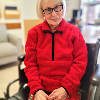 An elderly woman wearing a red fleece and glasses sits in a wheelchair indoors.