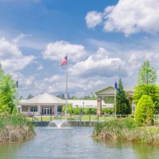 A scenic view of a building and fountain with a flagpole, surrounded by trees and a pond under a cloudy sky.