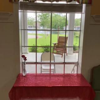 A view through a window displaying a porch with chairs and a table adorned with a single flower vase.