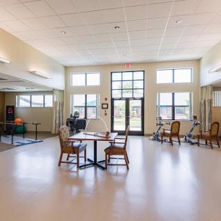 A spacious rehabilitation center featuring a bright community area with tables, chairs, and exercise equipment.