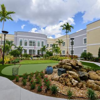 A modern building surrounded by a landscaped garden featuring palm trees and a stone decoration under a partly cloudy sky.