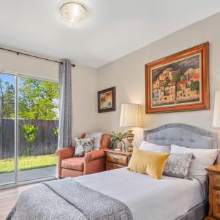 A cozy bedroom with a neatly made bed, an armchair by the window, and decorative paintings on the walls.