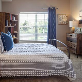 A cozy bedroom interior featuring a bed with patterned bedding, a wooden bookshelf, and a dresser beside a large window with blue curtains.