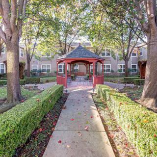 A charming garden walkway leads to a red gazebo, surrounded by neatly trimmed hedges and tall trees.
