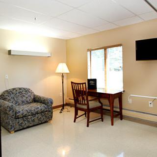 A cozy room with a sofa, wooden table and chair, lamp, and a TV, featuring a window that provides natural light.