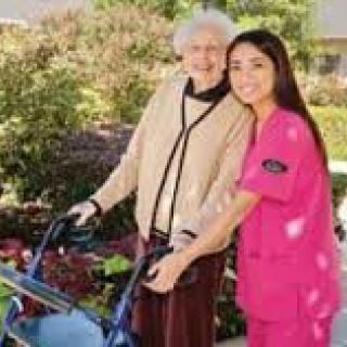 A caregiver in pink scrubs assists an elderly woman with a walker outdoors.