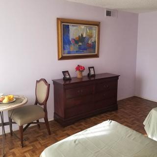 A cozy bedroom with a wooden dresser, a small round table, and a chair. The walls are adorned with a painting and decorated in soft tones.
