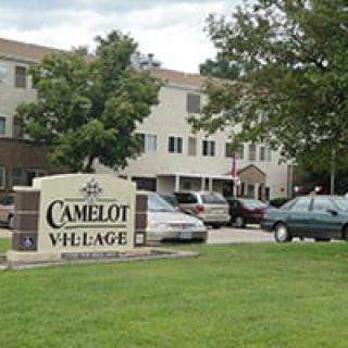 A view of the Camelot Village apartment complex with cars parked in front and trees lining the area.
