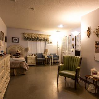 A cozy bedroom with a bed, dresser, and chairs, warmly lit by soft lighting.