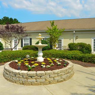 A beautifully landscaped garden with a central fountain surrounded by flowers and shrubs in front of a building.