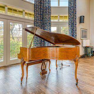 A beautifully crafted grand piano sits elegantly in a sunlit living room with large windows and hardwood floors.