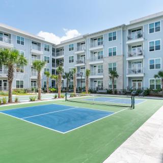 A modern apartment complex with an outdoor tennis court and palm trees under a clear blue sky.