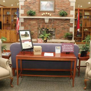 A cozy library lounge area with a sofa, chairs, and a wooden table in front of a brick fireplace, surrounded by bookshelves and decorative plants.