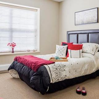 A cozy bedroom with a neatly made bed, featuring red and white decor accents, and a large window with blinds.