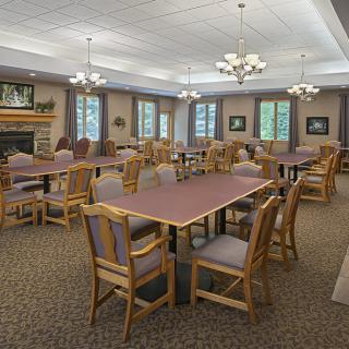 A spacious dining room with multiple tables and chairs, well-lit by chandeliers and natural light from the windows.