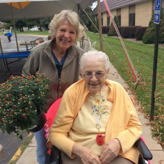 Two women, one elderly in a wheelchair, smile outdoors near a potted plant.