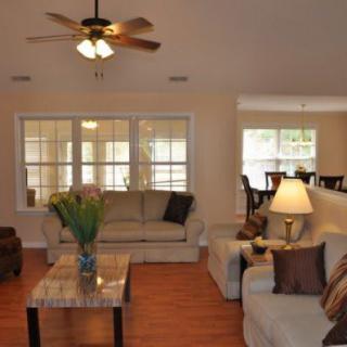 A cozy living room with a sofa, table, and lamp under a ceiling fan, featuring large windows.