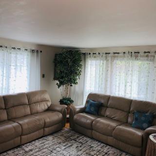 A cozy living room with two brown sofas, a patterned rug, and large windows adorned with white curtains.