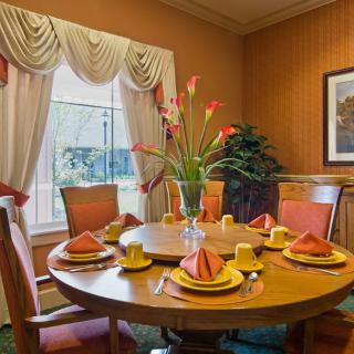 A well-decorated dining room featuring a round wooden table set with orange napkins and yellow dishes, complemented by floral arrangements and elegant curtains.