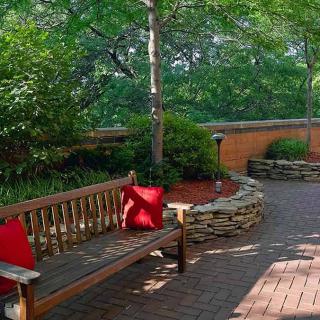 A peaceful garden setting with a wooden bench adorned with red cushions, surrounded by lush greenery and a stone wall.