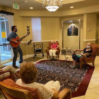A musician plays guitar for an audience of elderly people in a cozy room.