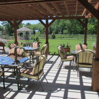 A sunny outdoor patio with tables and chairs under a pergola, overlooking a garden.