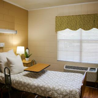 A cozy hospital room featuring a bed with a tray table, a lamp, and natural light from the window.