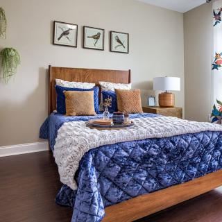 A cozy bedroom with a wooden bed frame, blue bedding, and decorative pillows. The room features artwork on the wall, a potted plant, and a stylish lamp.