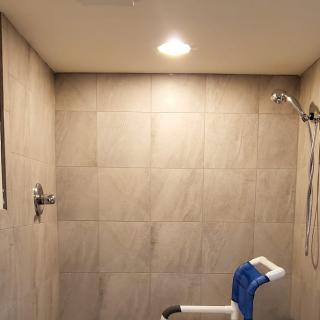 A tiled bathroom featuring a shower area with a shower chair under bright lighting.