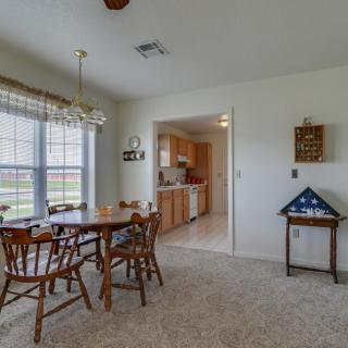 A cozy dining area with a wooden table and chairs, adjacent to a kitchen, featuring large windows and carpet flooring.