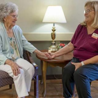 An elderly person and a caregiver hold hands while sitting together indoors.