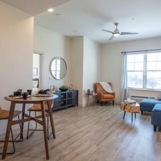 A modern living room with wood flooring, featuring a small dining table and chairs, cozy seating, and ample natural light from large windows.