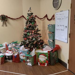 A festive office corner with a decorated Christmas tree surrounded by wrapped gifts and a whiteboard displaying event information.