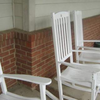 White rocking chairs on a porch with a brick wall backdrop.