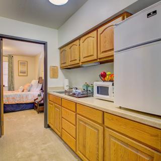 A compact kitchen area with wooden cabinets, a refrigerator, and a microwave, leading into a bedroom.