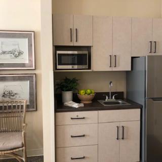 A compact modern kitchen with light cabinetry, featuring a microwave, refrigerator, and a small sink area.
