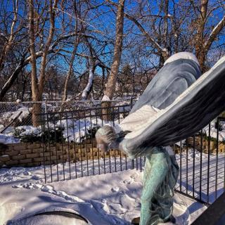 An angel statue covered in snow stands in a wintery outdoor setting with trees and a clear blue sky.