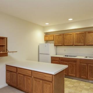A spacious kitchen with wooden cabinets and a white countertop, featuring a refrigerator and overhead lighting.