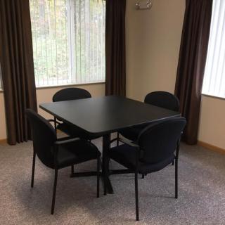 A simple meeting area featuring a black table with four chairs in an empty room with large windows.
