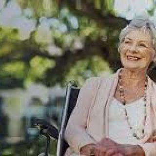 An elderly woman smiling while sitting in a wheelchair outdoors.