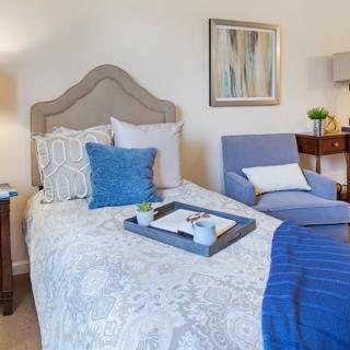 A cozy and well-decorated bedroom featuring a bed with patterned bedding, a blue throw blanket, and cushions. A tray is placed on the bed, and there is a comfortable chair along with a side table topped with a lamp.