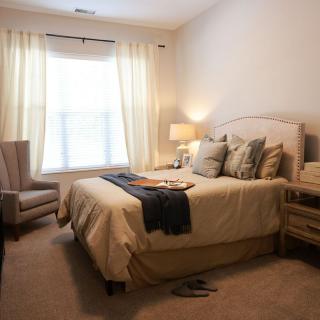 A cozy bedroom with a neatly made bed, soft lighting from a bedside lamp, and natural light streaming through the window curtains.