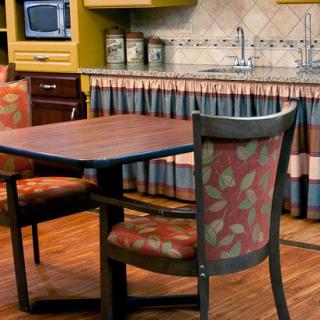 A cozy kitchen dining area featuring a wooden table and patterned chairs with a microwave and sink in the background.