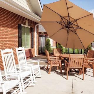 A cozy patio setup featuring white rocking chairs, a wooden table with matching chairs, and a large umbrella against a brick wall backdrop.