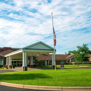 A building with an entrance and a flagpole stands against a partly cloudy sky, surrounded by a well-maintained lawn.