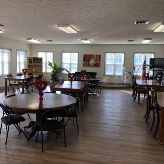 A spacious dining area with round tables, each adorned with red vases and surrounded by chairs, illuminated by natural light from large windows.